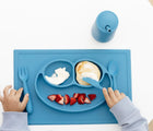 Child using ezpz Happy Utensils Set of 3 in blue together with ezpz mat and cup. Available from tenlittle.com